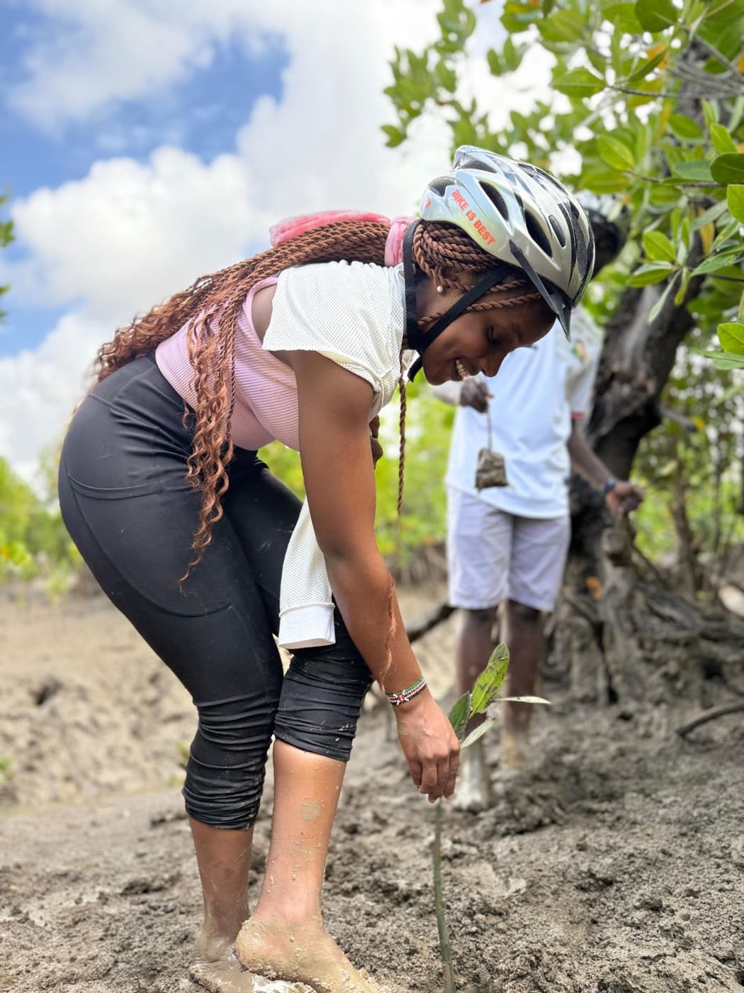 A rider in a hi-vis vest kneeling in the mud planting a mangrove seedling