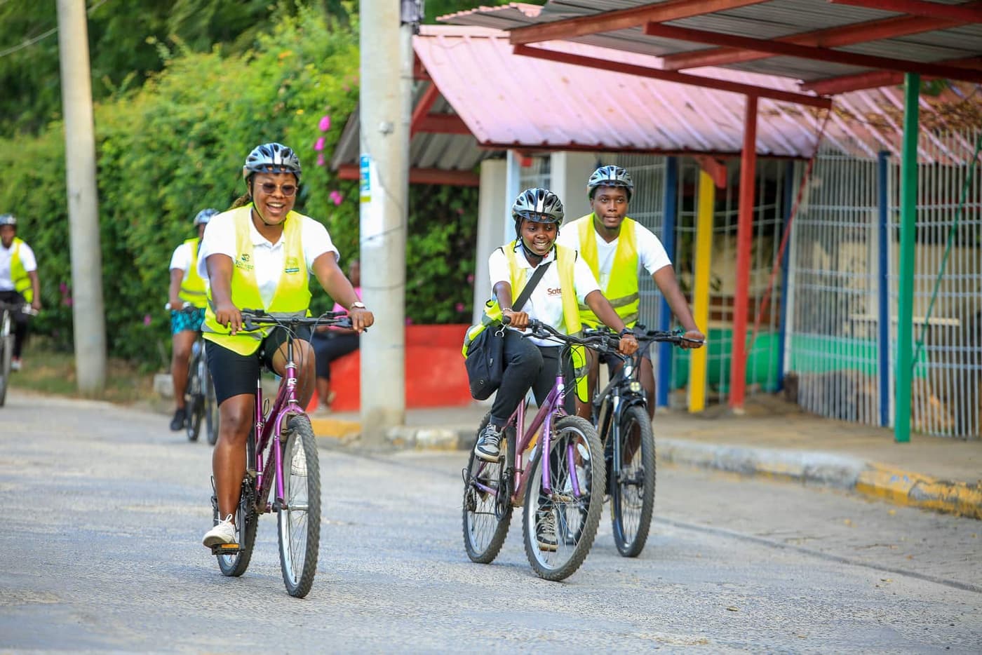 Three riders in hi-vis vests laughing as they ride past colourful shopfronts