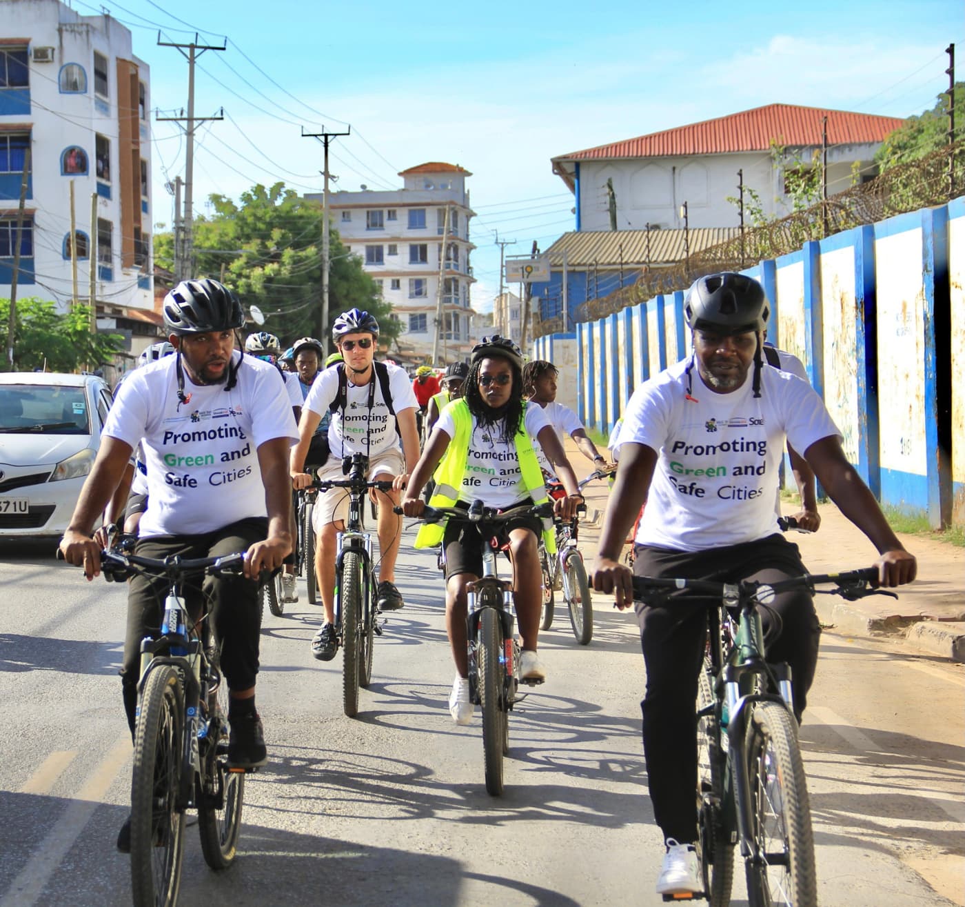 A group of riders in 'Promoting Green and Safe Cities' shirts cycling down a Mombasa road