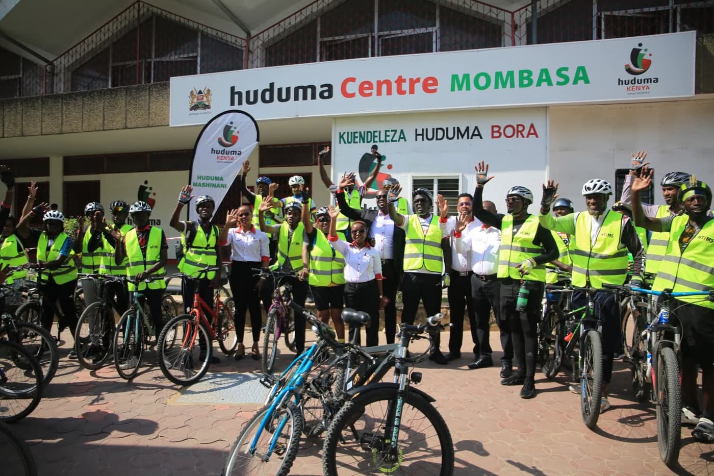 A large group of riders in hi-vis vests with arms raised in front of the Huduma Centre Mombasa banner