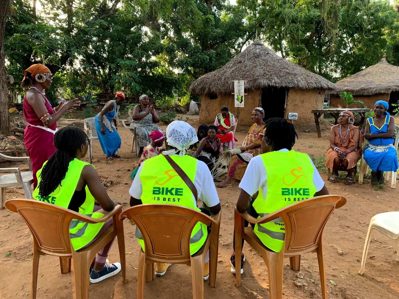 Riders in Bike is Best vests seated with village women in traditional dress beside thatched huts